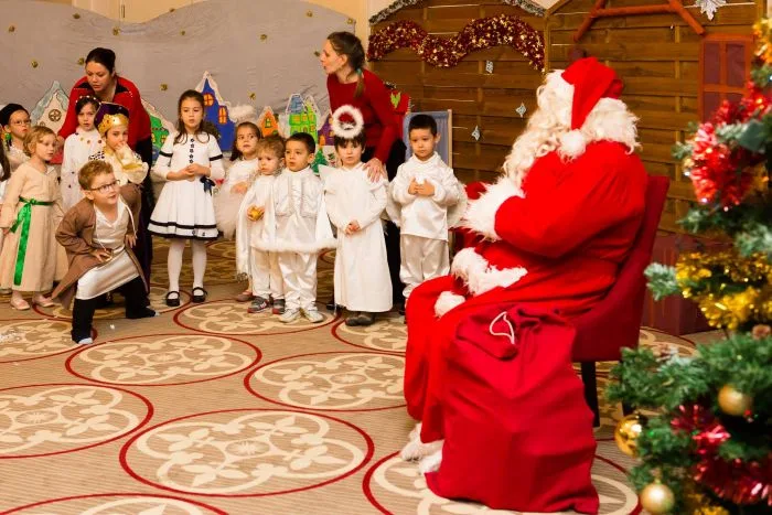 Moș Crăciun așezat în fața unui grup de copii costumați, la serbarea de grădiniță